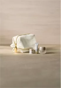 Ein luxuriöses weißes Kosmetiktäschchen mit goldenem Reißverschluss neben drei Hautpflegeproben auf einer grauen Marmoroberfläche bei dezenter Beleuchtung
